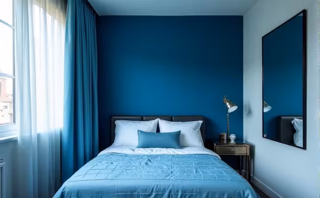 A small, modern bedroom showcasing a monochromatic color scheme. The room features various shades of blue, from a deep sapphire accent wall to a lighter sky-blue bedding and curtains. The ceiling is painted a shade lighter than the walls, adding an illusion of height. The room has a reflective, metallic side table and a large mirror on one wall, enhancing the overall space and brightness